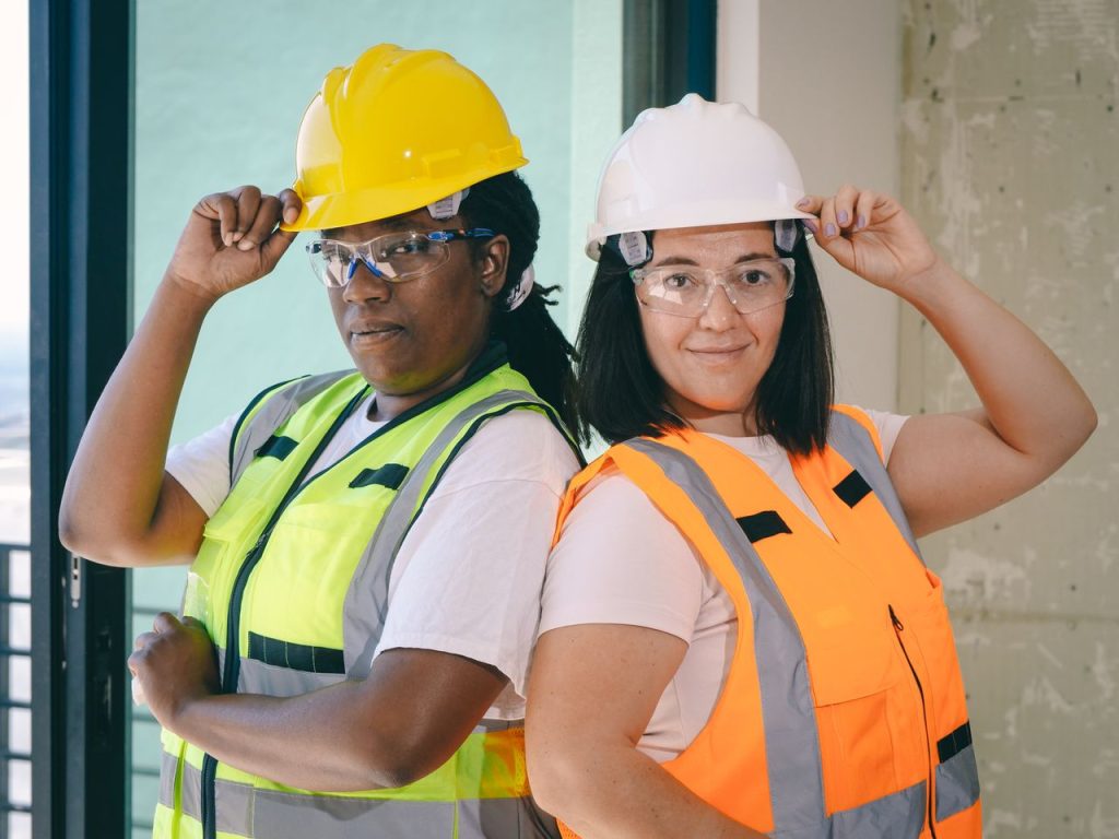 Two female construction workers wearing hard hats and safety gear, standing confidently indoors.